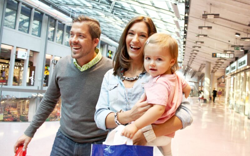 Ein Mann, eine Frau und ein kleines Kind mit Einkaufstaschen gehen gemeinsam durch ein hell erleuchtetes Einkaufszentrum.