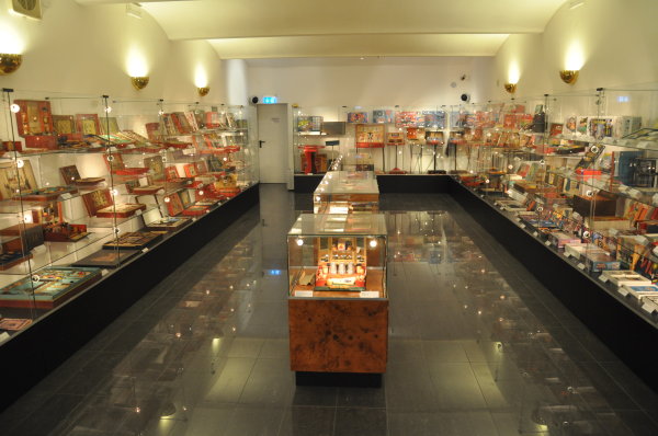 Ein gut beleuchteter Museumsraum mit Glasvitrinen, in denen verschiedene Vintage-Artikel, hauptsächlich kleine Schachteln und Sammlerstücke, ordentlich an den Wänden und in der Mitte angeordnet sind.