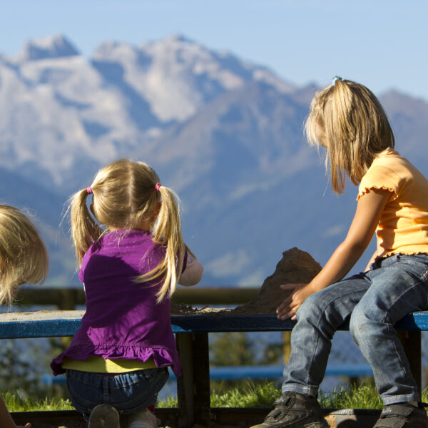 Drei kleine Kinder spielen auf einer Bank im Freien mit einer Bergkette im Hintergrund unter einem klaren blauen Himmel.