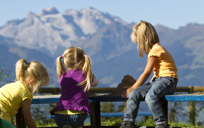 Drei kleine Kinder spielen auf einer Bank im Freien mit einer Bergkette im Hintergrund unter einem klaren blauen Himmel.