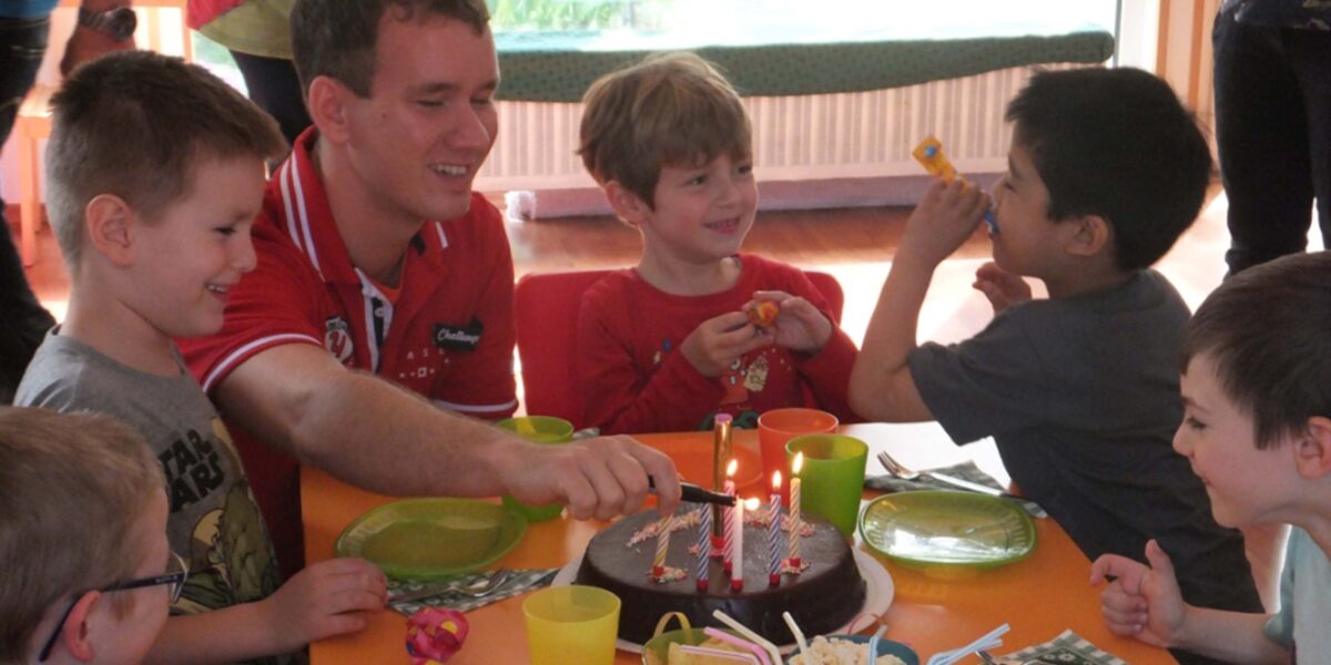Eine Gruppe kleiner Kinder und ein Erwachsener sitzen um einen Tisch mit Schokoladengeburtstagstorte, brennenden Kerzen und Partyzubehör herum und feiern drinnen.