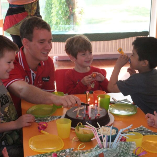 Eine Gruppe kleiner Kinder und ein Erwachsener sitzen um einen Tisch mit Schokoladengeburtstagstorte, brennenden Kerzen und Partyzubehör herum und feiern drinnen.