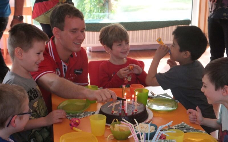 Eine Gruppe kleiner Kinder und ein Erwachsener sitzen um einen Tisch mit Schokoladengeburtstagstorte, brennenden Kerzen und Partyzubehör herum und feiern drinnen.