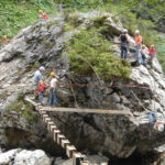 Menschen mit Helmen klettern und laufen auf Holzbrettern und Seilen, die an einem großen Felsen befestigt sind, in einem bewaldeten Outdoor-Abenteuerparcours.