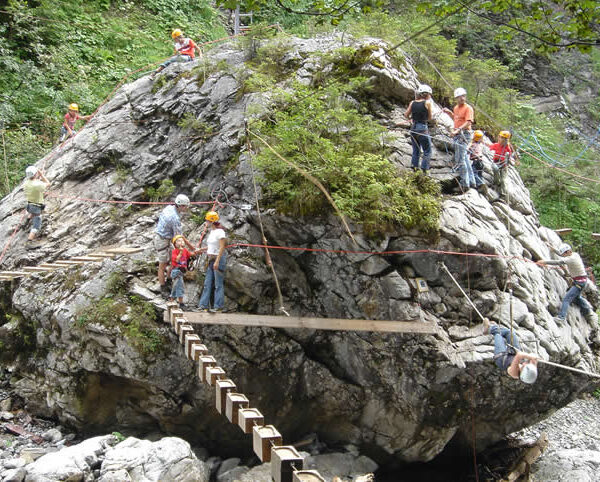 Menschen mit Helmen klettern und laufen auf Holzbrettern und Seilen, die an einem großen Felsen befestigt sind, in einem bewaldeten Outdoor-Abenteuerparcours.