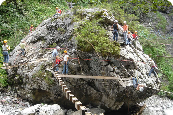 Menschen mit Helmen klettern und laufen auf Holzbrettern und Seilen, die an einem großen Felsen befestigt sind, in einem bewaldeten Outdoor-Abenteuerparcours.