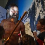 Ein Museumsführer spricht mit Kindern neben dem lebensgroßen Modell eines prähistorischen Menschen, der einen Stab hält, vor einer Bergkulisse.