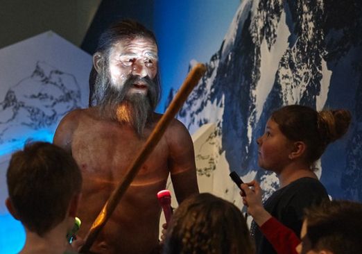 Ein Museumsführer spricht mit Kindern neben dem lebensgroßen Modell eines prähistorischen Menschen, der einen Stab hält, vor einer Bergkulisse.