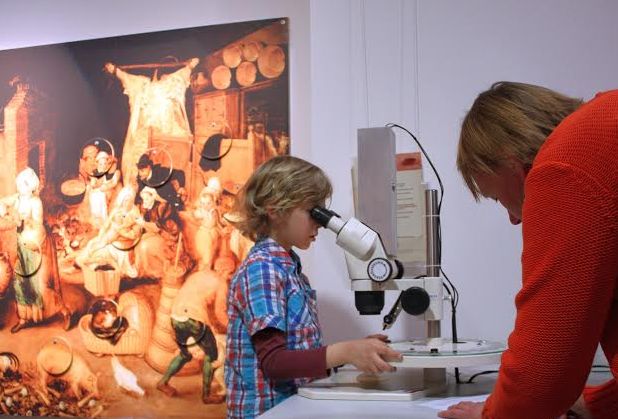 Ein Kind schaut in ein Mikroskop, während ein Erwachsener zusieht. Im Hintergrund ist an der Wand ein buntes Gemälde zu sehen.