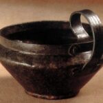 Eine dunkle Metallschale mit breitem Rand und einem dicken, geschwungenen Henkel an einer Seite, die auf einer neutralen Fläche steht.