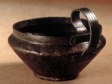 Eine dunkle Metallschale mit breitem Rand und einem dicken, geschwungenen Henkel an einer Seite, die auf einer neutralen Fläche steht.