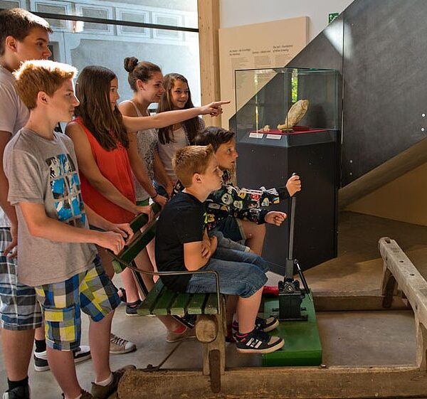 Eine Gruppe von Kindern und Jugendlichen schaut sich eine Museumsausstellung an. Zwei Jungen sitzen an einer interaktiven Ausstellung mit Hebeln.