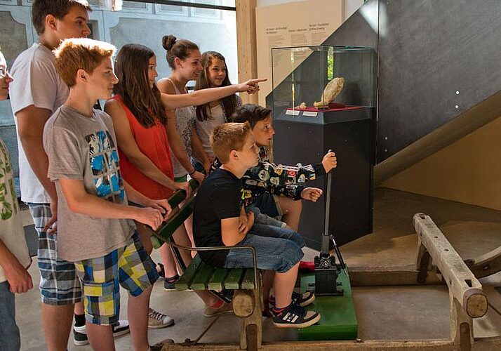 Eine Gruppe von Kindern und Jugendlichen schaut sich eine Museumsausstellung an. Zwei Jungen sitzen an einer interaktiven Ausstellung mit Hebeln.