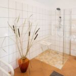 Ein gefliestes Badezimmer mit einer begehbaren Dusche, einer Glaswand, einem an der Wand befestigten Duschsitz, einer Topfpflanze, einem Regal mit gefalteten Handtüchern und einem weißen Waschbecken.