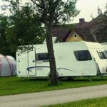 Zwei weiße Wohnwagen und ein graues Zelt sind auf einer Wiese in der Nähe eines Hauses geparkt, auf einem Wohnwagen sind Fahrräder montiert und ringsherum stehen Bäume.