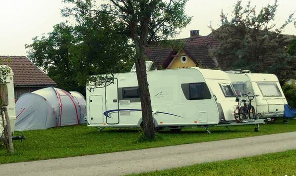Zwei weiße Wohnwagen und ein graues Zelt sind auf einer Wiese in der Nähe eines Hauses geparkt, auf einem Wohnwagen sind Fahrräder montiert und ringsherum stehen Bäume.