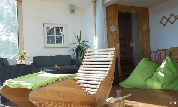 Ein sonnendurchfluteter Aufenthaltsraum mit einem hölzernen Lehnstuhl, grünen Sitzsäcken, einem Sofa, Topfpflanzen und einem kleinen Saunaraum im Hintergrund.