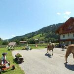 Kinder reiten auf Pferden und spielen an einem sonnigen Tag vor einem Gebäude im Chalet-Stil in einer bergigen, grasbewachsenen Gegend.