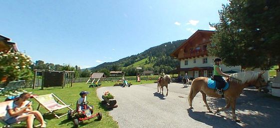 Kinder reiten auf Pferden und spielen an einem sonnigen Tag vor einem Gebäude im Chalet-Stil in einer bergigen, grasbewachsenen Gegend.