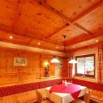 Holzgetäfeltes Esszimmer mit gepolsterten Sitzbänken, rot-weißer Tischdecke, Hängeleuchten, Fenster mit Vorhängen und gerahmter Wandkunst.