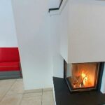 Ein kleiner Wohnbereich mit einem roten Sofa, einem hellen Couchtisch aus Holz und einem modernen weißen Kamin mit sichtbarem Feuer, daneben eine Küche mit Holzschränken.