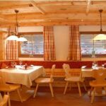 Ein gemütliches Restaurant mit Holzmöbeln, rot gemusterten Sitzpolstern, weißen Tischdecken und karierten Vorhängen, beleuchtet von Hängelampen.