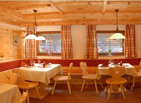 Ein gemütliches Restaurant mit Holzmöbeln, rot gemusterten Sitzpolstern, weißen Tischdecken und karierten Vorhängen, beleuchtet von Hängelampen.