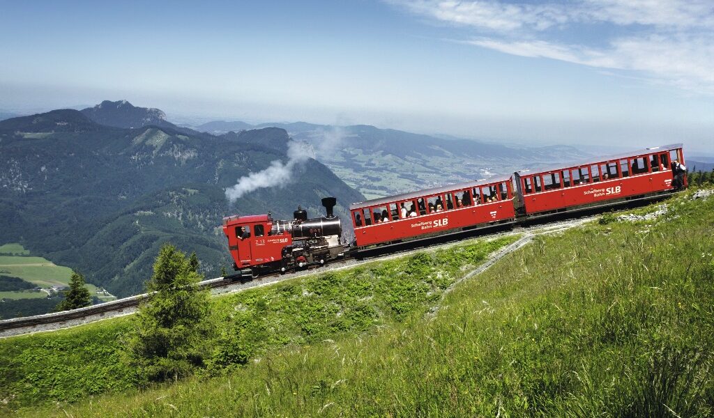 Ein roter Dampfzug fährt mit seinen Fahrgästen einen grasbewachsenen Berghang hinauf, umgeben von fernen Hügeln und Tälern unter einem klaren Himmel.