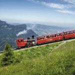 Ein roter Dampfzug fährt mit seinen Fahrgästen einen grasbewachsenen Berghang hinauf, umgeben von fernen Hügeln und Tälern unter einem klaren Himmel.