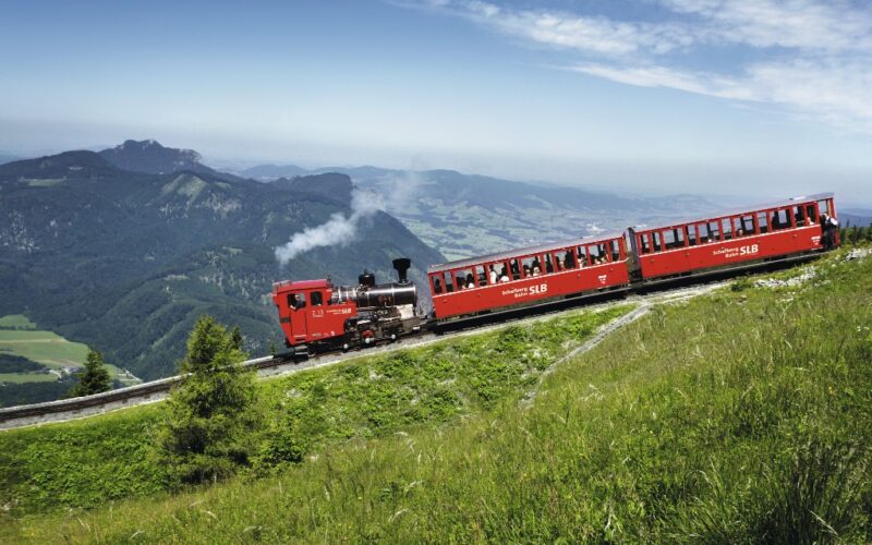 Ein roter Dampfzug fährt mit seinen Fahrgästen einen grasbewachsenen Berghang hinauf, umgeben von fernen Hügeln und Tälern unter einem klaren Himmel.