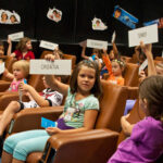 Die Kinder sitzen auf Stühlen im Hörsaal und halten Länderkarten mit Namen wie Kroatien, Türkei und Dänemark hoch, während sie scheinbar eine pädagogische Aktivität durchführen.