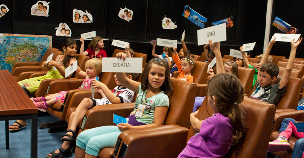 Die Kinder sitzen auf Stühlen im Hörsaal und halten Länderkarten mit Namen wie Kroatien, Türkei und Dänemark hoch, während sie scheinbar eine pädagogische Aktivität durchführen.