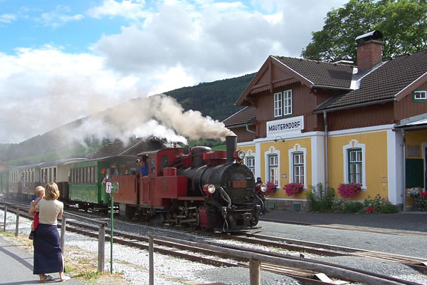 Ein roter Dampfzug kommt an einem kleinen Bahnhof mit der Aufschrift „Mauterndorf“ an. In der Nähe stehen Passagiere und eine Frau mit zwei Kindern und schauen zu.