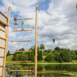 Mehrere Personen nehmen an einem Hochseilgarten und einer Seilrutsche im Freien in der Nähe eines Sees teil, umgeben von Bäumen und unter einem teilweise bewölkten Himmel.