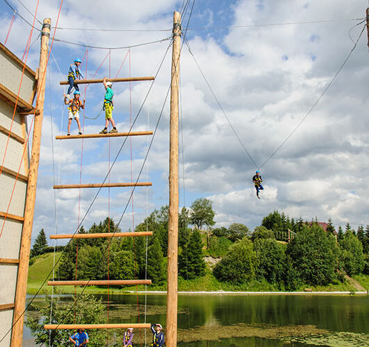 Mehrere Personen nehmen an einem Hochseilgarten und einer Seilrutsche im Freien in der Nähe eines Sees teil, umgeben von Bäumen und unter einem teilweise bewölkten Himmel.