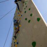 Zwei Personen mit Helmen und Gurten klettern unter einem klaren blauen Himmel an einer Kletterwand im Freien mit grünen und gelben Griffen.