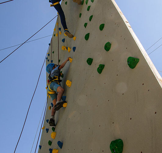Zwei Personen mit Helmen und Gurten klettern unter einem klaren blauen Himmel an einer Kletterwand im Freien mit grünen und gelben Griffen.