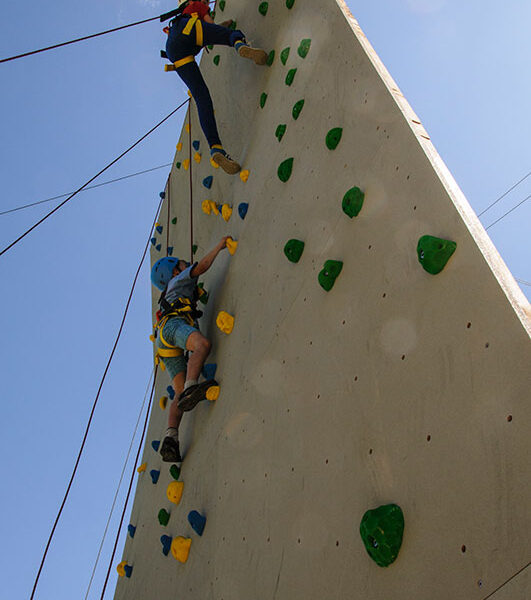 Zwei Personen mit Helmen und Gurten klettern unter einem klaren blauen Himmel an einer Kletterwand im Freien mit grünen und gelben Griffen.