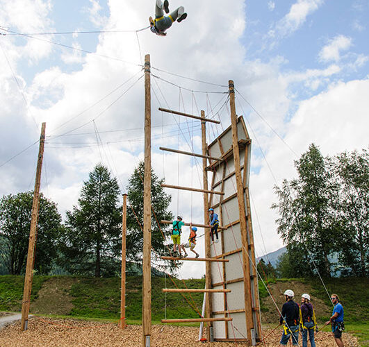 Eine Gruppe von Menschen nimmt an einem Hochseilgarten im Freien teil; eine Person hängt kopfüber in der Luft über der Struktur, während die anderen vom Boden aus zuschauen.