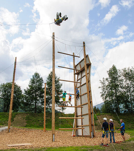 Eine Gruppe von Menschen nimmt an einem Hochseilgarten im Freien teil; eine Person hängt kopfüber in der Luft über der Struktur, während die anderen vom Boden aus zuschauen.