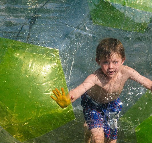 Ein kleiner Junge in Badehose läuft in einem großen aufblasbaren Plastikball auf dem Wasser und drückt seine Hände gegen die transparente Oberfläche.