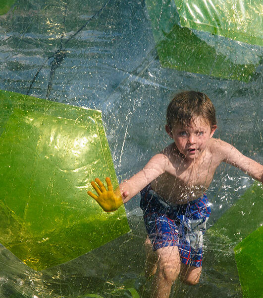 Ein kleiner Junge in Badehose läuft in einem großen aufblasbaren Plastikball auf dem Wasser und drückt seine Hände gegen die transparente Oberfläche.