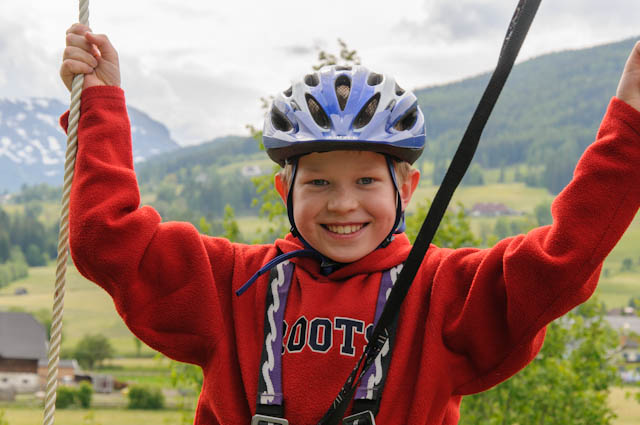 Lächelndes Kind mit Helm und rotem Pullover hält sich im Freien an Seilen fest, im Hintergrund grüne Hügel und Berge.