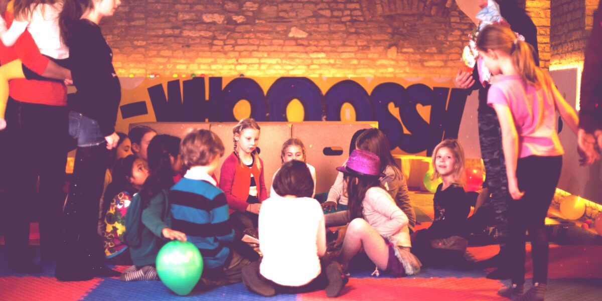 Eine Gruppe von Kindern und ein paar Erwachsene versammeln sich in einem Spielzimmer mit bunten Matten, Luftballons und einem "WHOOSH"-Schild im Hintergrund.