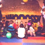 Eine Gruppe von Kindern und ein paar Erwachsene versammeln sich in einem Spielzimmer mit bunten Matten, Luftballons und einem "WHOOSH"-Schild im Hintergrund.