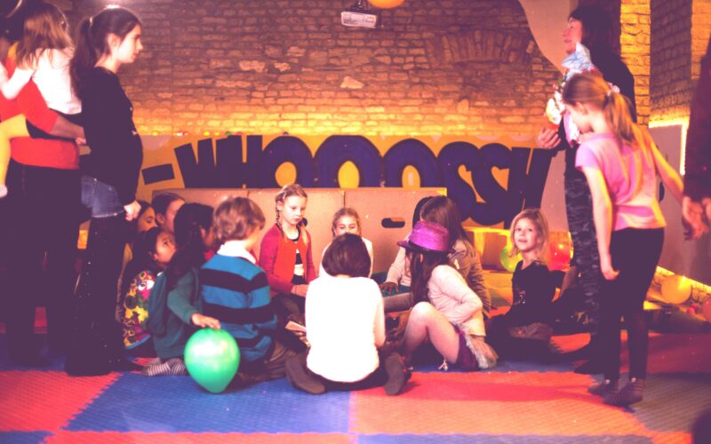 Eine Gruppe von Kindern und ein paar Erwachsene versammeln sich in einem Spielzimmer mit bunten Matten, Luftballons und einem "WHOOSH"-Schild im Hintergrund.