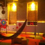 Innenspielbereich mit Backsteinwänden, weichem, farbenfrohem Bodenbelag, einem hängenden Netz, runden Schaumstoffblöcken und einem Zielbrett, beleuchtet von warmem, gelbem Licht.