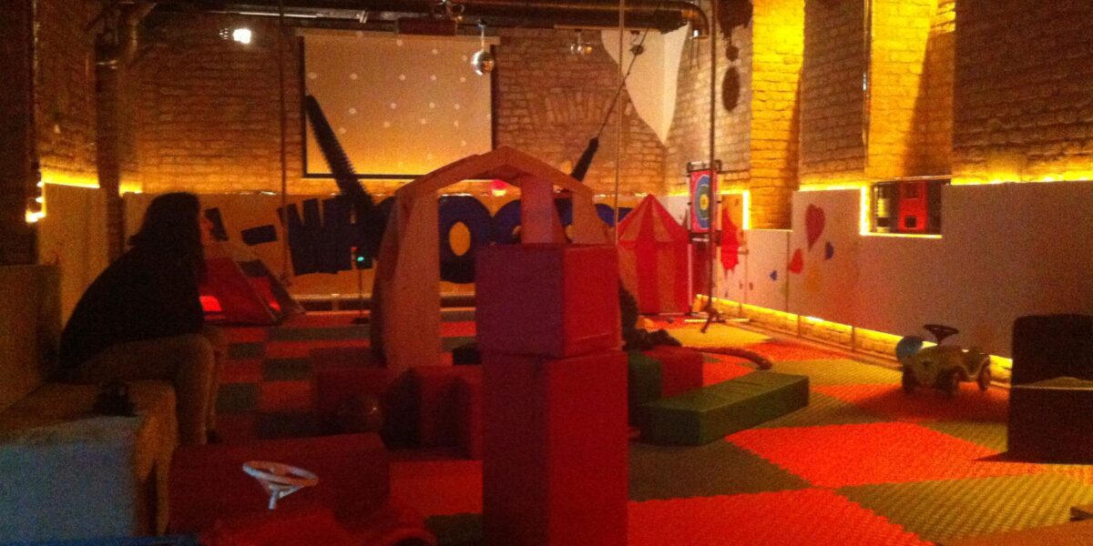 Spielzimmer für Kinder mit Schaumstoffmatten, Bauklötzen, Spielzeugfahrzeugen, Spielhäusern und gedämpfter Beleuchtung. Eine Person sitzt links auf einer Bank.
