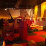 Spielzimmer für Kinder mit Schaumstoffmatten, Bauklötzen, Spielzeugfahrzeugen, Spielhäusern und gedämpfter Beleuchtung. Eine Person sitzt links auf einer Bank.