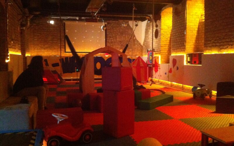 Spielzimmer für Kinder mit Schaumstoffmatten, Bauklötzen, Spielzeugfahrzeugen, Spielhäusern und gedämpfter Beleuchtung. Eine Person sitzt links auf einer Bank.